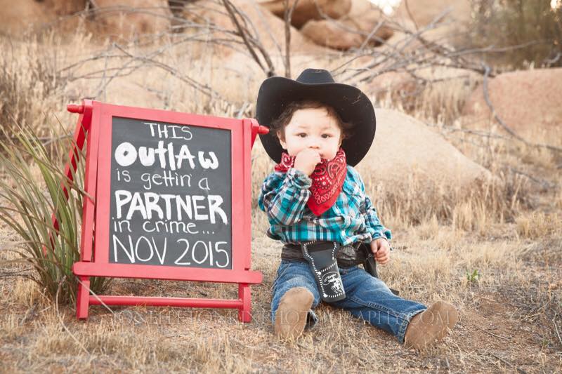 picture of a baby dressed in cowboy gear: blue flannel shirt, black cowboy hat, brown infant cowboy booties, red handkerchief, black belt, and gun holster. Next to sign that says "this outlaw is gettin' a partner in crime" for a fun a pregnancy announcement