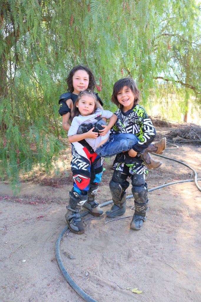 3 siblings in red and bright green dirt bike riding gear. the 2 brothers are holding up their sister in a princess carry while smiling. 